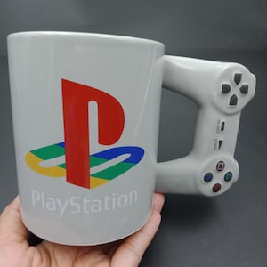 Op de afbeelding: Een witte keramische mok met het PlayStation-logo in rood, blauw, groen en geel. De handgreep is ontworpen als een PlayStation-controller, compleet met knoppen en een richtingskruis. Het woord "PlayStation" staat eronder gedrukt.