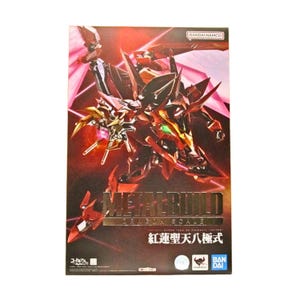 May include: A rectangular product box featuring a dynamic illustration of a red and gold mechanical robot figure. The text reads METAL BUILD DRAGON SCALE and includes the Bandai Namco logo. The background is a dark red gradient with light streaks.