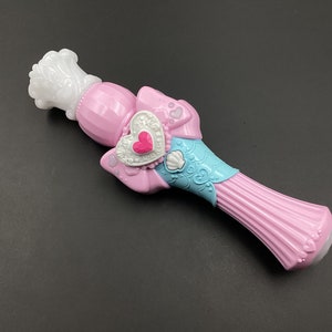 Op de afbeelding: Een roze en blauwe plastic toverstaf met een witte kroon bovenop. De toverstaf heeft een hartvormige hanger met een roze edelsteen in het midden. De toverstaf is versierd met een blauwe strik en een schelp.