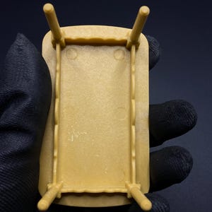 May include: A small, tan-colored plastic table with four legs. The table has a rectangular top with a decorative edge. The legs are cylindrical and attached to the corners of the table. The table is held in a gloved hand.