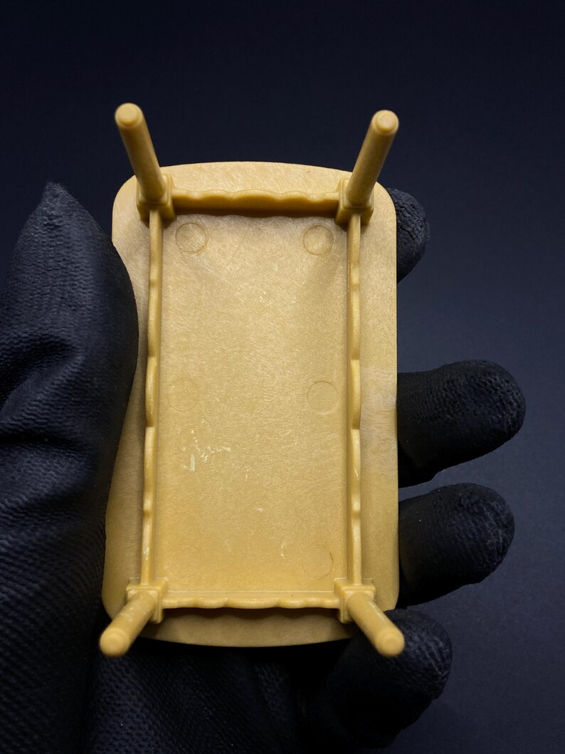 May include: A small, tan-colored plastic table with four legs. The table has a rectangular top with a decorative edge. The legs are cylindrical and attached to the corners of the table. The table is held in a gloved hand.