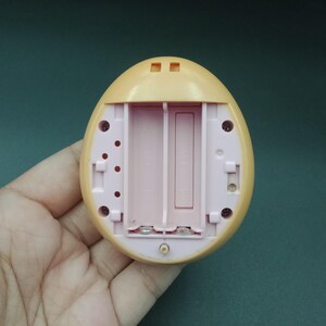 May include: A light yellow, egg-shaped electronic toy with a pink interior, featuring a battery compartment designed for two AA batteries. The toy has small circular and rectangular openings on its surface.