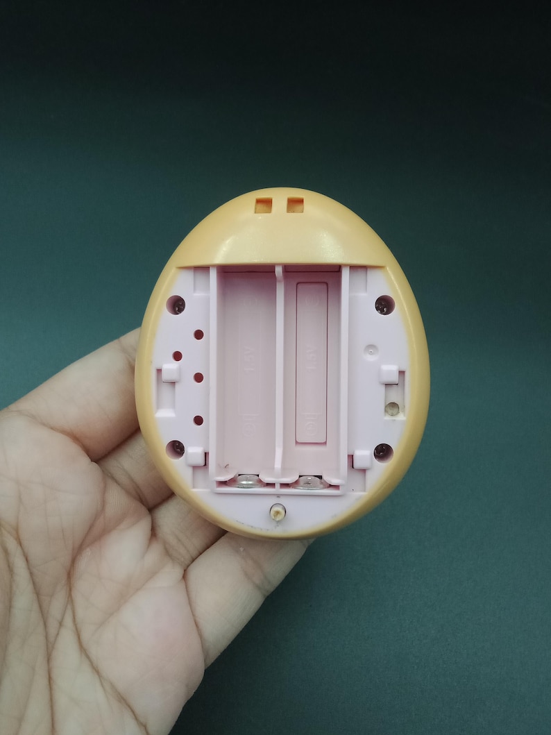 May include: A light yellow, egg-shaped electronic toy with a pink interior, featuring a battery compartment designed for two AA batteries. The toy has small circular and rectangular openings on its surface.