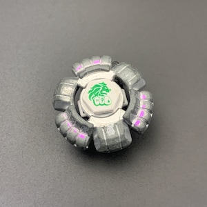 Puede incluir: Una peonza de metal plateado con una pieza central hexagonal blanca que muestra un gráfico de un león verde. El juguete tiene un anillo exterior segmentado en forma de engranaje con seis secciones distintas y reflejos morados.