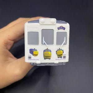 Puede incluir: Un tren de juguete blanco con techo gris y tres caras de tren de dibujos animados en la parte delantera. El tren tiene tres ventanas y limpiaparabrisas. El texto "WAKAYAMA ELECTRIC RAILWAY" está impreso en la parte inferior del tren.