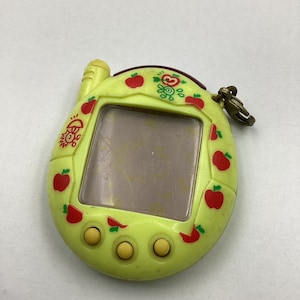 May include: A yellow digital pet toy with a red apple design. The toy has a small screen and three buttons. It has a small antenna on top and a loop for attaching a keychain.
