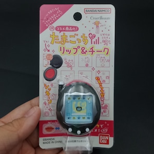 Op de afbeelding: Een zwart en rood Tamagotchi-thema lip- en cheek cosmeticacompact. De verpakking is roze en wit met Japanse tekst en illustraties. Het compact is in een hand gehouden.