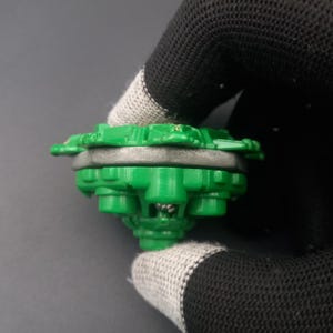 May include: A green and gray toy top with a multi-layered design. The top has a circular shape with a series of green protrusions around the edge. The center is a lighter gray color. The toy is held by a black and gray glove.