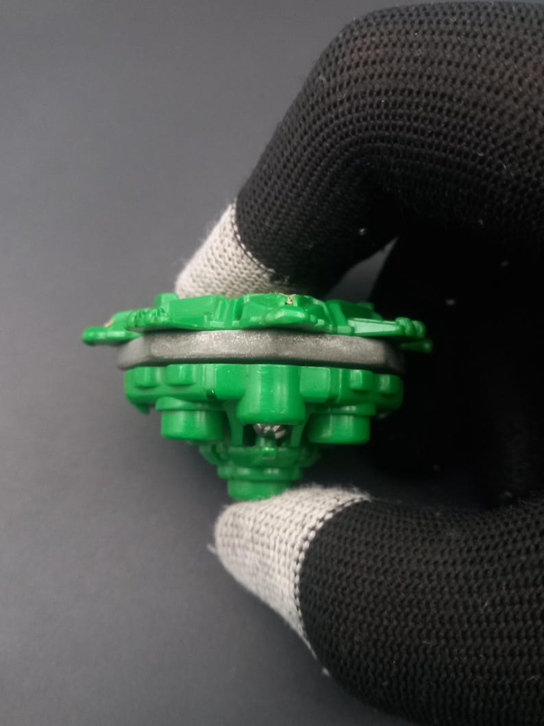 May include: A green and gray toy top with a multi-layered design. The top has a circular shape with a series of green protrusions around the edge. The center is a lighter gray color. The toy is held by a black and gray glove.