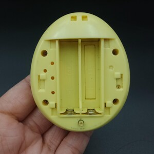 May include: A yellow, egg-shaped electronic device with an open battery compartment. The compartment is designed to hold two AA batteries. The device has several small holes and a circular indentation.