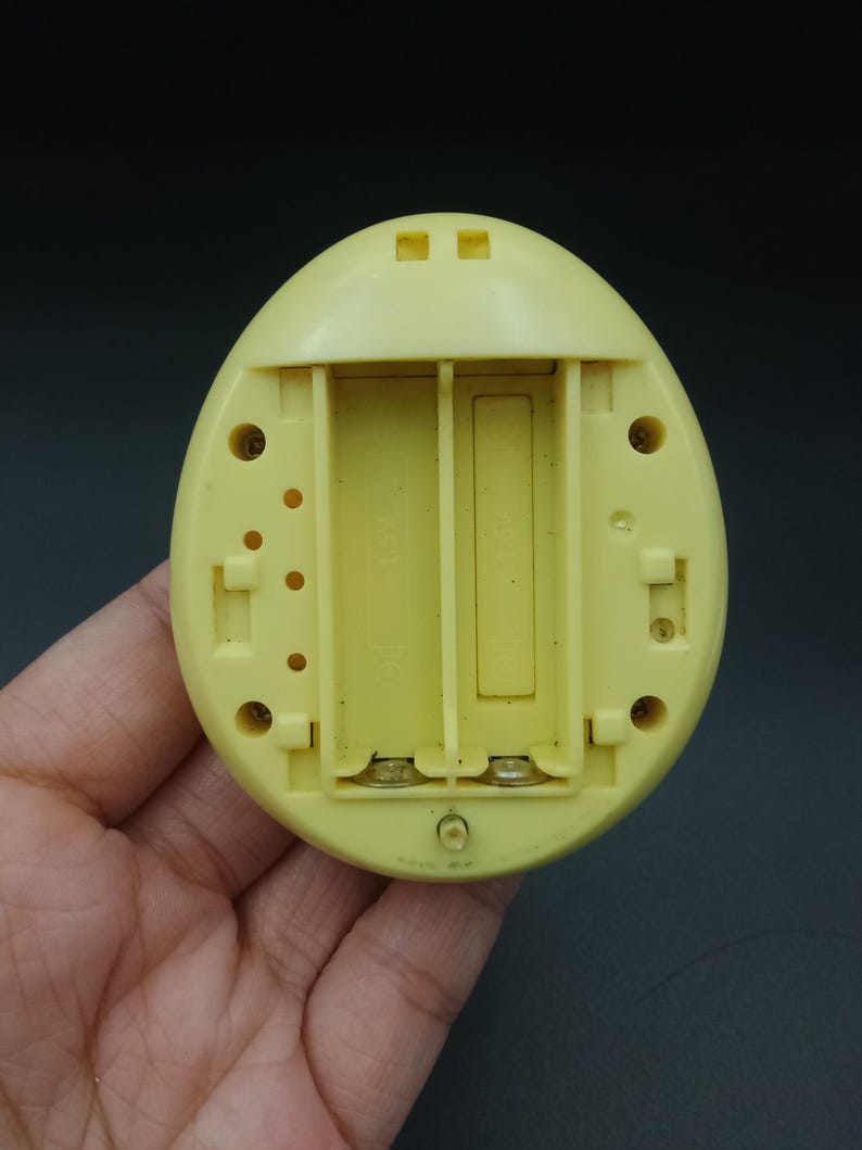 May include: A yellow, egg-shaped electronic device with an open battery compartment. The compartment is designed to hold two AA batteries. The device has several small holes and a circular indentation.
