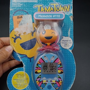 May include: A new, unopened Tamagotchi Town Memetchi #110 electronic pet toy in its original packaging. The packaging is blue with colorful graphics and text. The toy features a digital screen and a small orange character.