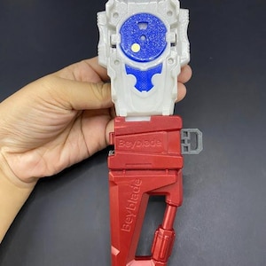 May include: A red, white, and blue Beyblade launcher. The launcher has a red handle with the word "Beyblade" printed on it. The top portion is white with a blue center and a blue pull cord.