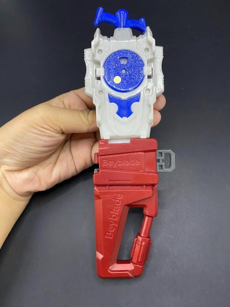 May include: A red, white, and blue Beyblade launcher. The launcher has a red handle with the word "Beyblade" printed on it. The top portion is white with a blue center and a blue pull cord.