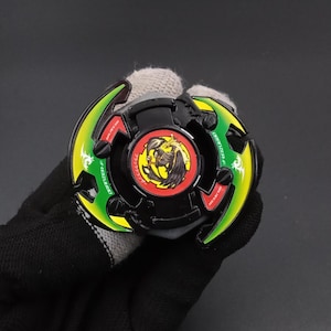 May include: A black and green spinning top toy with a dragon graphic in the center. The toy has green and yellow accents and red text. The toy is held in a gloved hand.