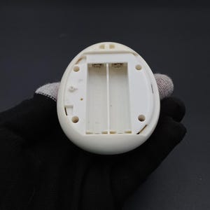 Puede incluir: Un dispositivo electr&oacute;nico blanco en forma de huevo con un compartimento de bater&iacute;a abierto. El compartimento est&aacute; dise&ntilde;ado para contener dos pilas AA. El dispositivo tiene un exterior liso y redondeado y varios agujeros peque&ntilde;os. El dispositivo es sostenido por una mano enguantada.