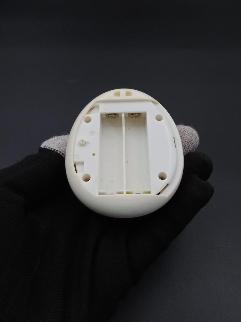 Puede incluir: Un dispositivo electr&oacute;nico blanco en forma de huevo con un compartimento de bater&iacute;a abierto. El compartimento est&aacute; dise&ntilde;ado para contener dos pilas AA. El dispositivo tiene un exterior liso y redondeado y varios agujeros peque&ntilde;os. El dispositivo es sostenido por una mano enguantada.