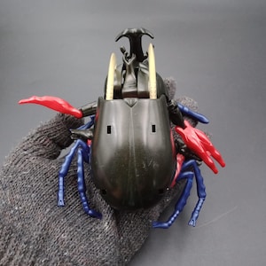 May include: A black and red toy beetle with blue legs and gold-colored horns. The toy has a segmented body and is held in a gray glove. The beetle's red claws and blue legs contrast with its dark body.