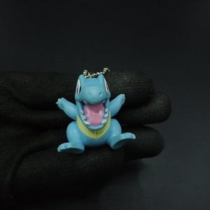 Puede incluir: Pequeña figura azul de Totodile con la boca abierta, revelando el interior rosa. La figura tiene una banda amarilla alrededor de la sección media y los brazos extendidos. Un llavero plateado está unido a la parte superior de la figura.