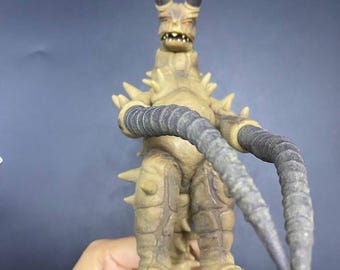 Può includere: Una action figure di mostro color marrone chiaro con grandi corna ricurve, una schiena a spillo e lunghe braccia segmentate. La figura ha un design dettagliato e strutturato ed è in posizione eretta.