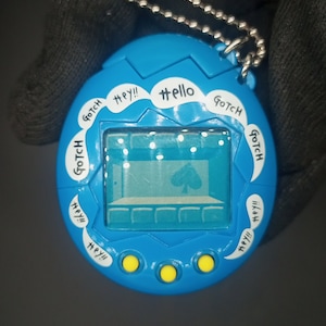 Puede incluir: Un juguete digital azul para mascotas con una pantalla que muestra una planta verde. El juguete tiene tres botones amarillos y el texto "GOTCH Hey!! #ello GOTCH Hey!! Hey!! GOTCH GOTCH Hey!! Hey!!" alrededor del borde.