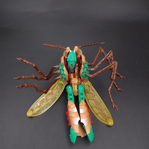 May include: A green, gold, and brown insect-like action figure with articulated limbs and wings. The toy has a detailed design, with visible segments and textures, and is posed against a dark background.