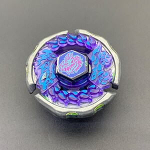 Könnte beinhalten: Ein kreisförmiges Kreiselspielzeug mit einem äußeren Ring aus silberfarbenem Metall und einem inneren Bereich aus durchscheinendem Violett. In der Mitte befindet sich ein sechseckiges Emblem mit einer pink-blauen Drachengrafik und violetten Details.