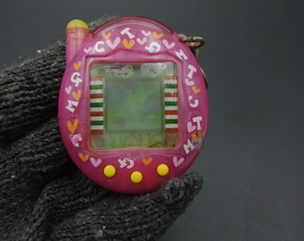 May include: A pink handheld digital pet toy with a small screen and buttons. The toy is decorated with hearts and letters. The screen displays a pixelated image. A yellow antenna is on top. A metal keychain is attached.
