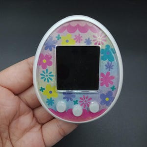 Puede incluir: Un juguete digital en forma de huevo con un marco blanco y un diseño floral. La pantalla es negra y hay tres botones grises debajo. El patrón floral incluye flores rosas, amarillas, azules y moradas. El dispositivo se sostiene en una mano.