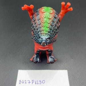 May include: A vibrant monster figurine with a red body, arms, and feet. The head is a mix of green, black, and orange, with a textured pattern. The figurine has a menacing expression and the text "B627 P6250" on a white label.