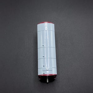 Könnte beinhalten: Ein weißer zylindrischer Behälter mit roter Ober- und Unterseite. Der Behälter hat vier horizontale Linien und eine kleine rechteckige Öffnung an der Vorderseite.