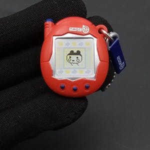 May include: A red handheld digital pet game with a silver frame around the screen. The screen displays a pixelated character. The game has three blue buttons and a blue and silver keychain. The text "TMGC" is visible on the game and keychain.