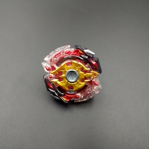 Puede incluir: Un trompo de plástico rojo, dorado y transparente. El juguete tiene un diseño circular con múltiples capas y un disco central dorado. El trompo presenta detalles en rojo y negro y está diseñado para el juego competitivo.