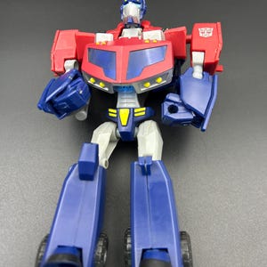 Könnte beinhalten: Eine rote, blaue und silberne Optimus Prime Actionfigur. Der Spielzeugroboter hat einen roten Oberkörper, blaue Beine und ein silbernes Gesicht. Die Figur hat ein gelb-schwarzes Emblem auf der Brust und schwarze Räder an den Füßen.