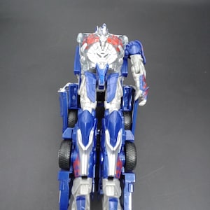 May include: A blue and silver action figure of a robot, with red accents on the chest and arms. The figure is in a standing position, with wheels visible on the lower body. The robot has a detailed design.