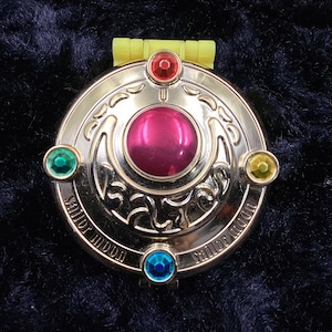 May include: A gold-coloured compact with a crescent moon design and five colourful gems. The compact reads "Sailor Moon" on the bottom.