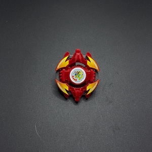 Könnte beinhalten: Rote und gelbe Plastik-Beyblade mit einem weißen und grünen Design in der Mitte. Die Beyblade hat vier Punkte und ist zum Drehen ausgelegt.