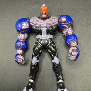 Op de afbeelding: Gedetailleerd actiefiguur van een robot met een rode kop, blauwe armen en zwarte benen. De robot heeft een zilveren borst en paarse accenten. De figuur staat rechtop met de armen langs de zijden. De armen en benen van de robot zijn gedetailleerd.
