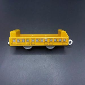 Puede incluir: Un vagón de tren de juguete amarillo con dos ruedas grises y un gancho blanco. El vagón tiene ventanas rectangulares con detalles grises y amarillos. La parte superior tiene dos secciones elevadas.