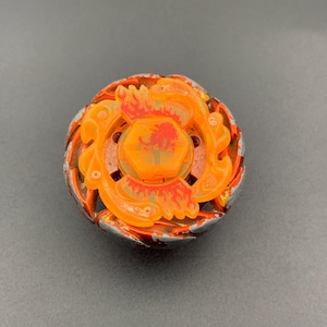 Può includere: Una trottola circolare caratterizzata da un centro in plastica arancione traslucida con un design a forma di fiamma e un tappo esagonale centrale. L'anello esterno è composto da elementi grigio metallizzato e rosso scuro frastagliati.