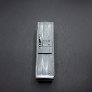 May include: A vintage silver-coloured lighter with a ribbed design. The lighter has a rectangular shape with a small switch and a black bottom. The lighter is on a dark grey surface.