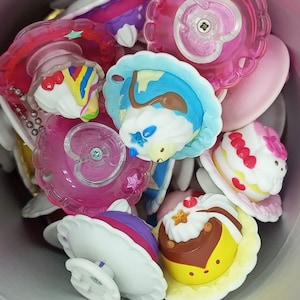 May include: A collection of colorful plastic toy charms shaped like different desserts, including cupcakes, cakes, and ice cream cones. The charms are decorated with glitter and other embellishments.