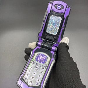 May include: A purple and black flip phone toy featuring a holographic screen panel, a numeric keypad with an 'ENTER' button, and a stylised emblem above the keys. The device has a mechanical design with visible screws and a circular detail at the top.