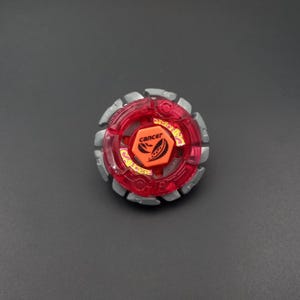 Pode incluir: Um brinquedo de pião circular com uma estrutura de plástico vermelho translúcido e um anel externo de metal prateado. O parafuso central laranja exibe o texto "cancer" e um gráfico de caranguejo, com texto "cancer" adicional no plástico vermelho.