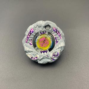 May include: A circular silver-colored spinning top toy featuring a central yellow hexagonal piece with a pink graphic design. The outer metallic frame has intricate sculpted details and recessed sections filled with a vibrant purple patterned inlay.