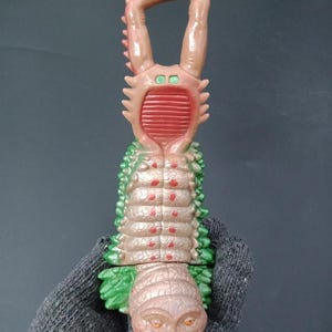 May include: A detailed, multi-colored toy creature with a segmented body, featuring shades of green, brown, and red. The toy has a unique design with large, curved appendages and a textured surface. The label at the bottom reads "B750P8010".