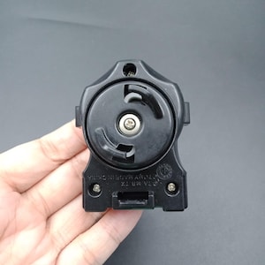 Könnte beinhalten: Eine schwarze Kunststoff-Spielzeugkomponente mit einem kreisförmigen Zentrum und einer rechteckigen Basis. Die Komponente hat eine Schraube in der Mitte und der Text "OTA MB TX" und "TOY MADE IN CHINA" ist sichtbar. Die Komponente wird in einer Hand gehalten.