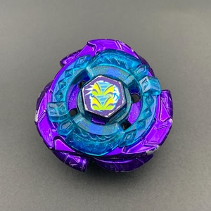 Op de afbeelding: Een paars en blauw tol-speeltje met een metallic paarse buitenring met geometrische patronen en een doorschijnende blauwe binnenlaag. Het zeshoekige middenstuk toont een geel en blauw grafisch ontwerp van een gestileerd wezengezicht.