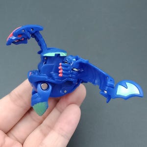 Może przedstawiać: Niebieska zabawka w kształcie stworzenia z czerwonymi i zielonymi akcentami. Zabawka ma smoczą głowę i skrzydła. Zabawka jest trzymana w dłoni.