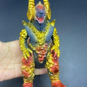 Puede incluir: Figura de monstruo colorida con un diseño detallado. La figura tiene una cabeza azul, boca roja y cuernos dorados. El cuerpo es una mezcla de oro, rojo y azul, con escamas texturizadas. El juguete está sostenido por una mano.